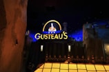 A nighttime scene featuring a brightly lit sign reading 'Gusteau's' glowing in yellow with blue accents. The sign includes an illuminated image of a chef holding a dish, surrounded by stars. The background includes dark blue sky with stars and buildings or structures in the foreground, giving the impression of an urban setting.