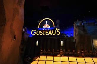 A nighttime scene featuring a brightly lit sign reading 'Gusteau's' glowing in yellow with blue accents. The sign includes an illuminated image of a chef holding a dish, surrounded by stars. The background includes dark blue sky with stars and buildings or structures in the foreground, giving the impression of an urban setting.