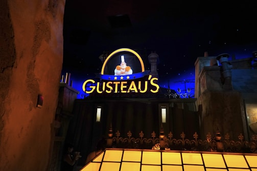 A nighttime scene featuring a brightly lit sign reading 'Gusteau's' glowing in yellow with blue accents. The sign includes an illuminated image of a chef holding a dish, surrounded by stars. The background includes dark blue sky with stars and buildings or structures in the foreground, giving the impression of an urban setting.