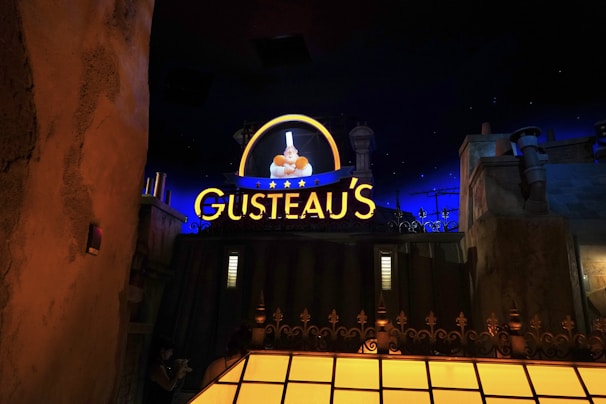 A nighttime scene featuring a brightly lit sign reading 'Gusteau's' glowing in yellow with blue accents. The sign includes an illuminated image of a chef holding a dish, surrounded by stars. The background includes dark blue sky with stars and buildings or structures in the foreground, giving the impression of an urban setting.