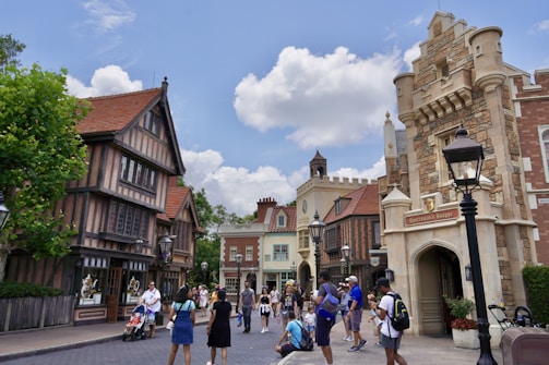 A lively street scene features a variety of tourists walking and exploring. The architecture is styled with medieval European elements, including stone facades, pointed roofs, and decorative wooden beams. Several people are taking photos, while others are pushing strollers or chatting. Trees and lampposts line the street under a bright, partly cloudy sky.