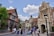 A lively street scene features a variety of tourists walking and exploring. The architecture is styled with medieval European elements, including stone facades, pointed roofs, and decorative wooden beams. Several people are taking photos, while others are pushing strollers or chatting. Trees and lampposts line the street under a bright, partly cloudy sky.