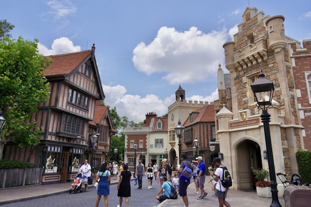 A lively street scene features a variety of tourists walking and exploring. The architecture is styled with medieval European elements, including stone facades, pointed roofs, and decorative wooden beams. Several people are taking photos, while others are pushing strollers or chatting. Trees and lampposts line the street under a bright, partly cloudy sky.