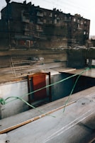 An empty wooden chair with a red backrest is placed in an indoor setting. A thin green wire is stretched across the foreground. A metal rack and other objects are placed on dusty shelves and countertops. The reflection of an urban building with several windows and cars parked outside is visible through a glass window.