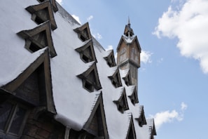 A steep roof with multiple triangular dormer windows covered in snow, alongside a tall clock tower with wooden detailing under a clear blue sky.