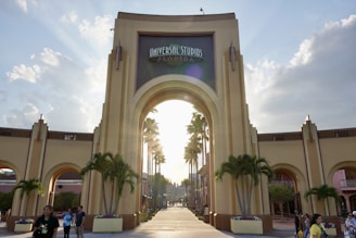 An iconic archway forms the entrance to a popular theme park with the sign 'Universal Studios Florida'. Tall palm trees line the walkway leading up to the entrance, and there are several small groups of people walking or standing around. The sky is partly cloudy with the sun setting or rising, casting a warm glow over the scene.