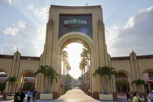 An iconic archway forms the entrance to a popular theme park with the sign 'Universal Studios Florida'. Tall palm trees line the walkway leading up to the entrance, and there are several small groups of people walking or standing around. The sky is partly cloudy with the sun setting or rising, casting a warm glow over the scene.