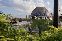 A large globe sculpture with the word 'UNIVERSAL' in bold letters is prominently displayed. The foreground features lush green foliage and purple flowers, with a body of water and a bridge visible behind the plants. The sky is partly cloudy, creating a bright and pleasant atmosphere.