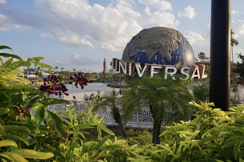 A large globe sculpture with the word 'UNIVERSAL' in bold letters is prominently displayed. The foreground features lush green foliage and purple flowers, with a body of water and a bridge visible behind the plants. The sky is partly cloudy, creating a bright and pleasant atmosphere.