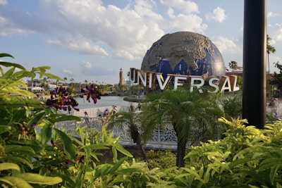A large globe sculpture with the word 'UNIVERSAL' in bold letters is prominently displayed. The foreground features lush green foliage and purple flowers, with a body of water and a bridge visible behind the plants. The sky is partly cloudy, creating a bright and pleasant atmosphere.