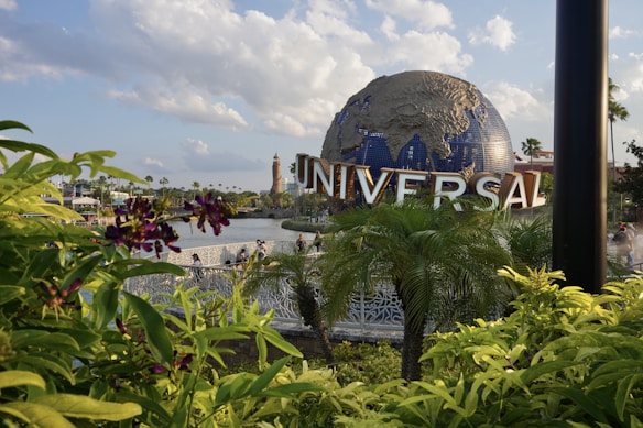 A large globe sculpture with the word 'UNIVERSAL' in bold letters is prominently displayed. The foreground features lush green foliage and purple flowers, with a body of water and a bridge visible behind the plants. The sky is partly cloudy, creating a bright and pleasant atmosphere.
