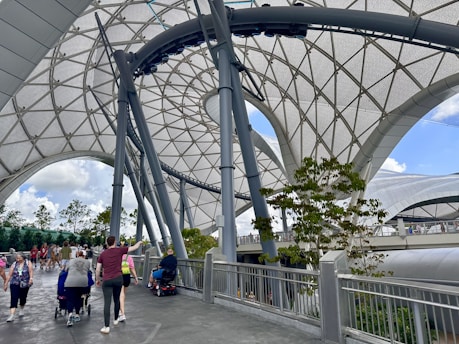 A covered walkway features large, geometric metal and fabric structures creating an open and airy atmosphere. People are walking and using mobility aids along the pathway, which has a modern and futuristic design. Trees and a partially cloudy sky are visible in the background.