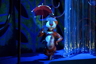 A colorful, cartoon-style animal character stands confidently holding a red umbrella. The character is covered in floral patterns and appears to be in a whimsical environment with shiny, curtain-like strands in the background. The lighting is primarily blue with some contrasting orange and purple tones.
