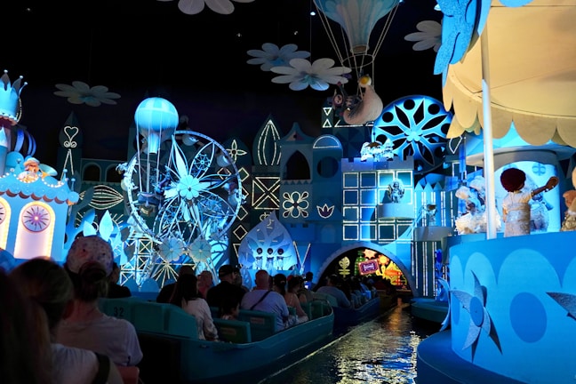 A whimsical amusement ride featuring a vibrant display of animated figures and colorful sets. The scene is dominated by shades of blue and white, with various characters, a Ferris wheel, and a hot air balloon. Visitors are seated in small boats as they enjoy the immersive experience.