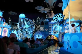 A whimsical amusement ride featuring a vibrant display of animated figures and colorful sets. The scene is dominated by shades of blue and white, with various characters, a Ferris wheel, and a hot air balloon. Visitors are seated in small boats as they enjoy the immersive experience.
