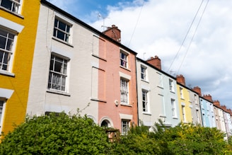 A row of colorful terrace houses with pastel-painted facades, featuring vibrant shades such as yellow, pink, and blue. Each house has multiple white-framed windows and brick chimneys, with greenery and bushes in the foreground and a partly cloudy sky above.