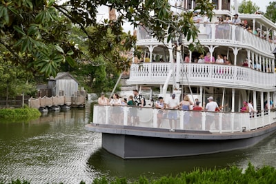 A large, multi-tiered riverboat filled with people is navigating a scenic waterway surrounded by lush greenery. The upper and lower decks of the boat feature ornate railings and are crowded with passengers enjoying the view. Trees overhang a portion of the water, and a rustic wooden structure can be seen near the shoreline.