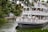 A large, multi-tiered riverboat filled with people is navigating a scenic waterway surrounded by lush greenery. The upper and lower decks of the boat feature ornate railings and are crowded with passengers enjoying the view. Trees overhang a portion of the water, and a rustic wooden structure can be seen near the shoreline.