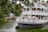 A large, multi-tiered riverboat filled with people is navigating a scenic waterway surrounded by lush greenery. The upper and lower decks of the boat feature ornate railings and are crowded with passengers enjoying the view. Trees overhang a portion of the water, and a rustic wooden structure can be seen near the shoreline.