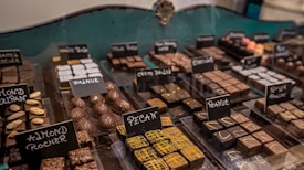 An assortment of chocolates displayed in a glass case. Each type of chocolate has a small sign indicating its flavor or filling, such as pecan, walnut, almond, and peanut. The chocolates are arranged in neat rows and vary in shape, size, and decoration.