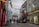 A bustling narrow street lined with colorful buildings and shops, adorned with various international flags and Irish flags. People are walking, shopping, and enjoying the atmosphere. The shops have vibrant signage and window displays, and there are flower baskets and bright banners adding to the festive environment.