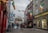 A bustling narrow street lined with colorful buildings and shops, adorned with various international flags and Irish flags. People are walking, shopping, and enjoying the atmosphere. The shops have vibrant signage and window displays, and there are flower baskets and bright banners adding to the festive environment.
