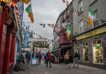A bustling narrow street lined with colorful buildings and shops, adorned with various international flags and Irish flags. People are walking, shopping, and enjoying the atmosphere. The shops have vibrant signage and window displays, and there are flower baskets and bright banners adding to the festive environment.