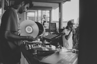 A black and white photo featuring a DJ holding a vinyl record and operating turntables, with sound equipment visible. A person in a cap is drinking from a can while standing beside the DJ. The setting appears to be an outdoor venue, as indicated by the open space and visible architectural elements like columns and windows.