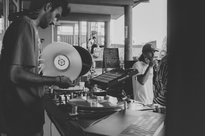 A black and white photo featuring a DJ holding a vinyl record and operating turntables, with sound equipment visible. A person in a cap is drinking from a can while standing beside the DJ. The setting appears to be an outdoor venue, as indicated by the open space and visible architectural elements like columns and windows.