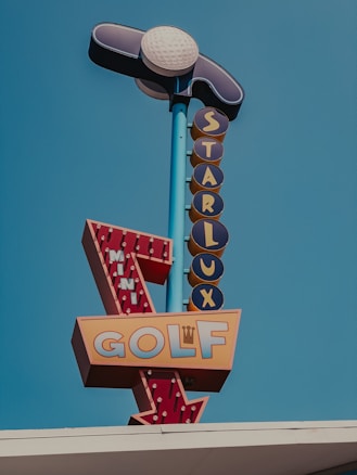 A colorful vintage-style sign with a golf ball and club at the top. The sign reads 'Starlux' vertically and 'Mini Golf' with an arrow pointing downwards. The background is a clear blue sky.