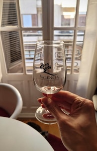A hand is holding a clear wine glass with the text 'Valencia Denominación de Origen' printed on it. The background shows a window with multiple panes and a view of buildings outside. Sunlight creates a warm ambiance, casting soft shadows.