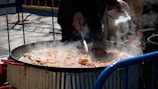 A large metal pan filled with various ingredients is being stirred by a person. Steam rises from the pan, indicating cooking in progress. The setting appears to be outdoors with visible blue railings and people nearby.