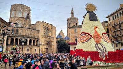 A bustling crowd gathers in a historic plaza, engaged in a festive event. The centerpiece is a large floral structure resembling a figure adorned with an ornate sun-like headdress, crafted from white and red flowers. Surrounding this are iconic Gothic cathedral buildings with intricate architecture, including towers and arches.