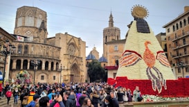 A bustling crowd gathers in a historic plaza, engaged in a festive event. The centerpiece is a large floral structure resembling a figure adorned with an ornate sun-like headdress, crafted from white and red flowers. Surrounding this are iconic Gothic cathedral buildings with intricate architecture, including towers and arches.