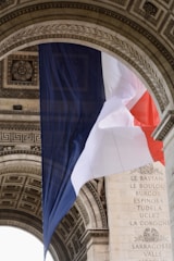 A large French flag is draped elegantly under an ornate stone archway. The flag's tricolor of blue, white, and red contrasts with the detailed carvings and inscriptions on the stone. The architectural elements feature intricate patterns and classical motifs.