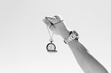 A hand holding up an antique pocket watch on a chain against a clear sky. The wrist wears a metallic wristwatch, suggesting a contrast between old and new timekeeping devices.