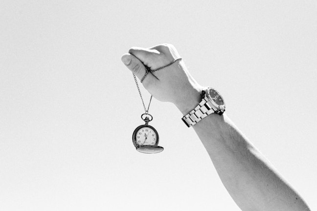 A hand holding up an antique pocket watch on a chain against a clear sky. The wrist wears a metallic wristwatch, suggesting a contrast between old and new timekeeping devices.