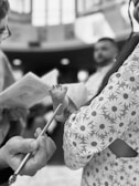 A close-up of a baby's foot being held up by someone wearing a patterned garment, possibly at a ceremony. A hand holding a thin metallic object, perhaps a religious or ceremonial tool, is also visible. Blurred figures in the background suggest an indoor setting with soft lighting.