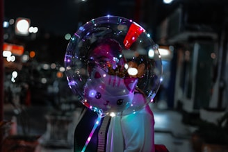 A transparent balloon with a cute cat face design and colorful LED lights inside. A person is partially visible behind the balloon, adding a mysterious and whimsical element. The background is an out-of-focus cityscape, possibly taken at night, with bokeh light effects.
