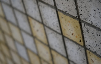 A grid of square tiles arranged in a diagonal pattern, some tiles are off-white while others are a light tan color. The tiles have a speckled texture with small dark spots scattered across their surfaces.