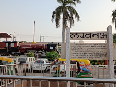 A large red and black train locomotive is displayed near a station platform with a palm tree to the right. Several vehicles, including autorickshaws and cars, are parked nearby. A prominent sign in Hindi is seen in the foreground. Rail tracks and power lines are visible in the background.