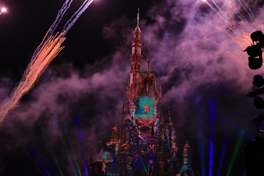 A nighttime scene featuring a brightly illuminated castle, surrounded by colorful fireworks with smoke in the sky. The castle is adorned with vibrant projections, enhancing its intricate architecture. The dark background contrasts with the light display, adding a sense of magic and celebration.