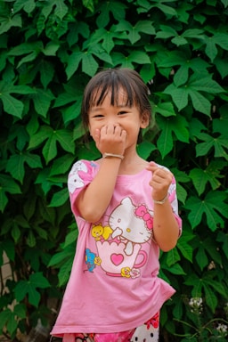 A young girl with a playful expression stands in front of a lush background of large green leaves. She is wearing a pink shirt featuring cartoon characters and is covering her mouth with one hand, possibly laughing.