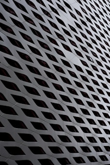 A close-up of a modern building facade featuring a repetitive pattern of diamond-shaped openings, creating an illusion of depth and movement. The structure appears to be made of metallic panels with a sleek, glossy finish.