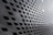 A modern architectural facade featuring a pattern of diamond-shaped openings arranged in a repetitive, grid-like structure. The sleek, metallic surface creates a futuristic and dynamic appearance with alternating light and shadow effects.