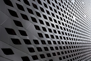 A modern architectural facade featuring a pattern of diamond-shaped openings arranged in a repetitive, grid-like structure. The sleek, metallic surface creates a futuristic and dynamic appearance with alternating light and shadow effects.
