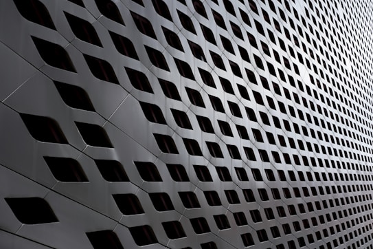 A modern architectural facade featuring a pattern of diamond-shaped openings arranged in a repetitive, grid-like structure. The sleek, metallic surface creates a futuristic and dynamic appearance with alternating light and shadow effects.