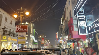 A bustling street scene featuring numerous shops and signs illuminated at night. People are walking or standing along the crowded sidewalks while many cars are parked or driving through the street. The storefronts display varied signage and lights, including names like 'Bag Boutique,' 'Bombay Dyeing,' 'Titan,' and 'Swayamvar.' Street lamps are lit, contributing to the overall illumination. Overhead, multiple wires crisscross the street.