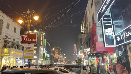 A bustling street scene featuring numerous shops and signs illuminated at night. People are walking or standing along the crowded sidewalks while many cars are parked or driving through the street. The storefronts display varied signage and lights, including names like 'Bag Boutique,' 'Bombay Dyeing,' 'Titan,' and 'Swayamvar.' Street lamps are lit, contributing to the overall illumination. Overhead, multiple wires crisscross the street.