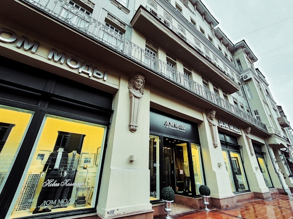 A stylish building with a fashion boutique featuring decorative architectural details. Mannequins displaying suits are visible through large, illuminated windows. The facade has ornamental columns and sculptural elements, with a sign in Cyrillic above the entrance.